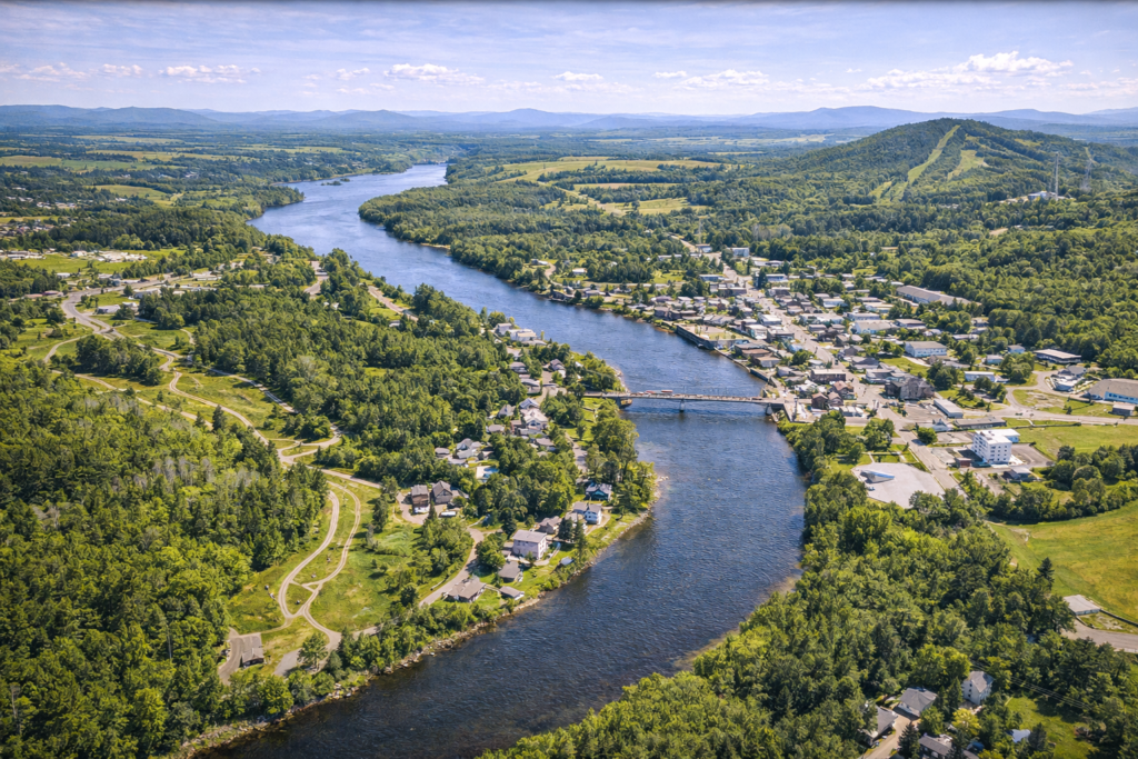 Aerial view of Fort Kent, Maine at the confluence of the St. John and Fish Rivers, showing downtown, forested hills, Lonesome Pine Trails, and vibrant summer landscape in Aroostook County.