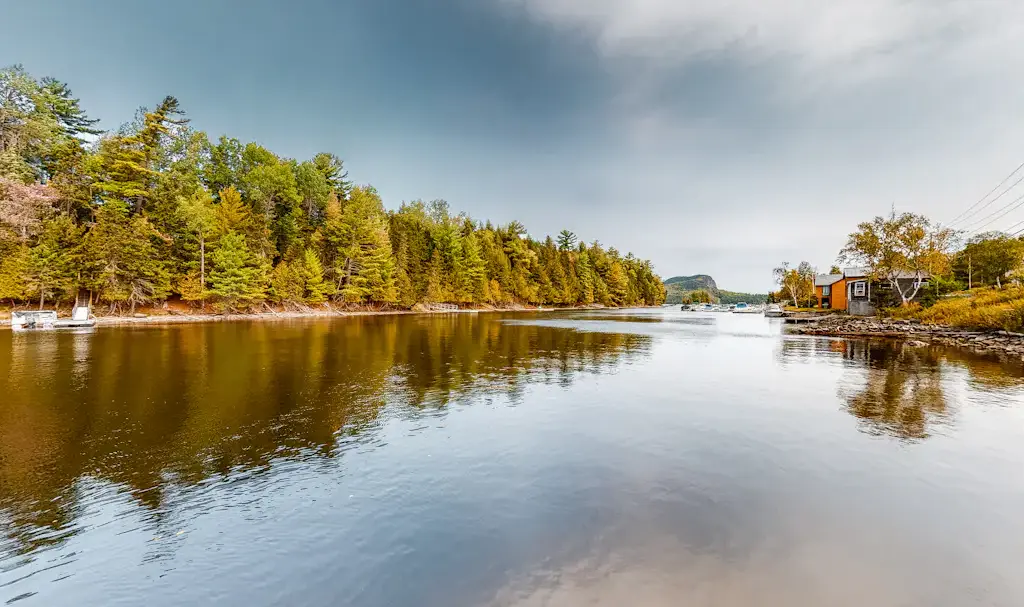 Get news on Moosehead News scenic views of the Moose River in Rockwood, Maine, featuring vibrant autumn foliage reflecting on calm water with rustic waterfront cabins and a distant view of Mount Kineo under a cloudy sky.