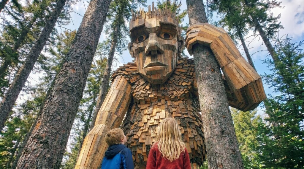 A young boy in a blue jacket and a young blonde girl in a red sweater stand looking up at a towering giant troll sculpture made of recycled wood scrap and shingles peering out from behind tall pine trees.