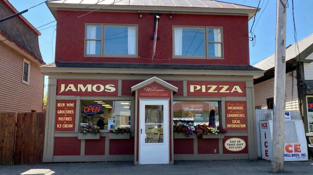 The exterior storefront of Jamos Pizza in downtown Greenville, Maine, featuring a red and tan facade, window flower boxes, and gold lettering advertising pizza and Dagwood sandwiches.